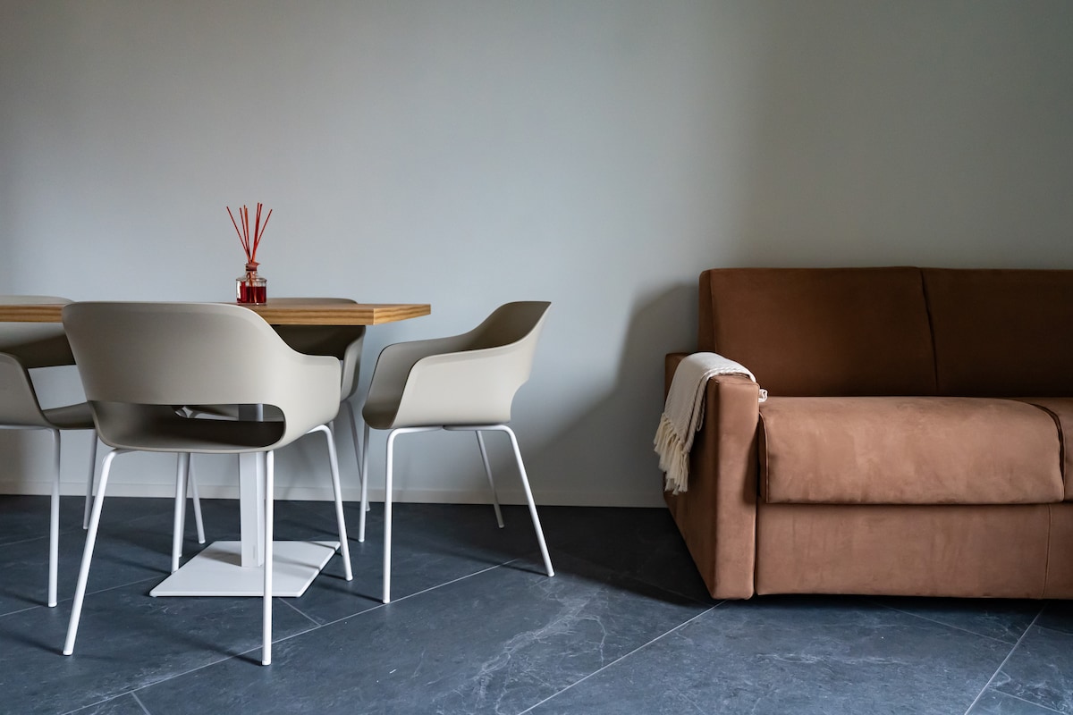 A dining area features a wooden table surrounded by four light-colored chairs. A brown sofa is positioned nearby, complemented by a cozy throw draped across its arm. The space is enhanced by gray tiled flooring and neutral-colored walls.