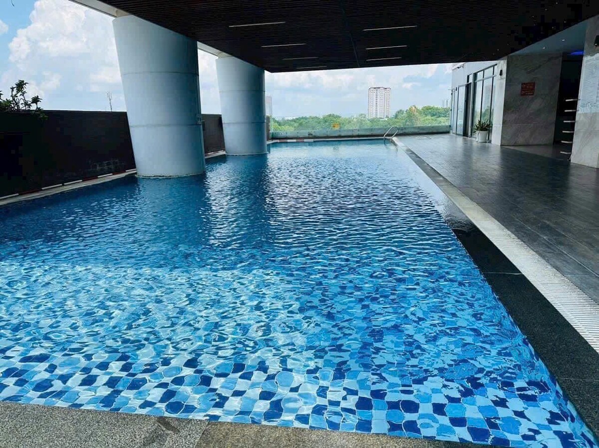 A shimmering pool is set under a covered area, showcasing clear blue water and a patterned tile border. Surrounding greenery is visible through expansive glass walls, creating a relaxed atmosphere. Sunlight reflects off the water's surface, enhancing the inviting look of the space.
