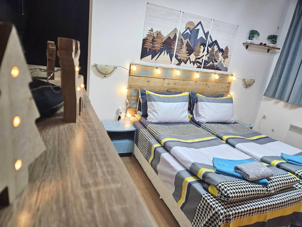A sleeping area features two single beds with a plaid duvet and decorative pillows, framed by a headboard made of wooden planks. Soft lighting from string lights adds a cozy touch. A wooden dresser with decorative accents is visible in the foreground.