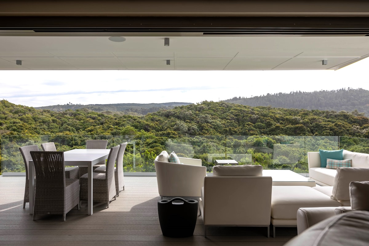 An outdoor patio features a stylish dining table accompanied by woven chairs, and a comfortable seating area with soft cushions. Retractable glass doors provide an unobstructed view of lush greenery, enhancing the sense of openness and connection to the surrounding nature.