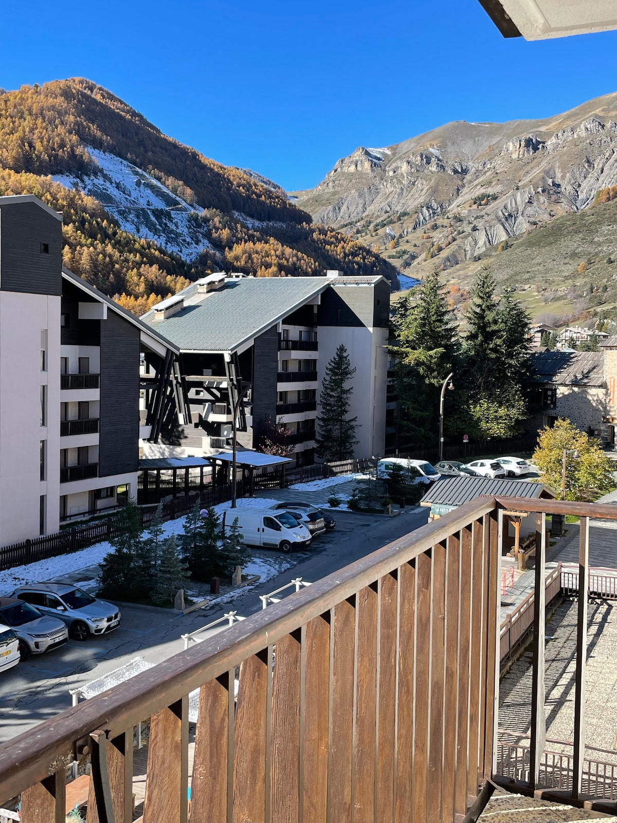 A view from the terrace showcases a landscape of mountains and buildings. The scene includes several parked vehicles surrounded by trees and autumn foliage, complemented by clear blue skies.