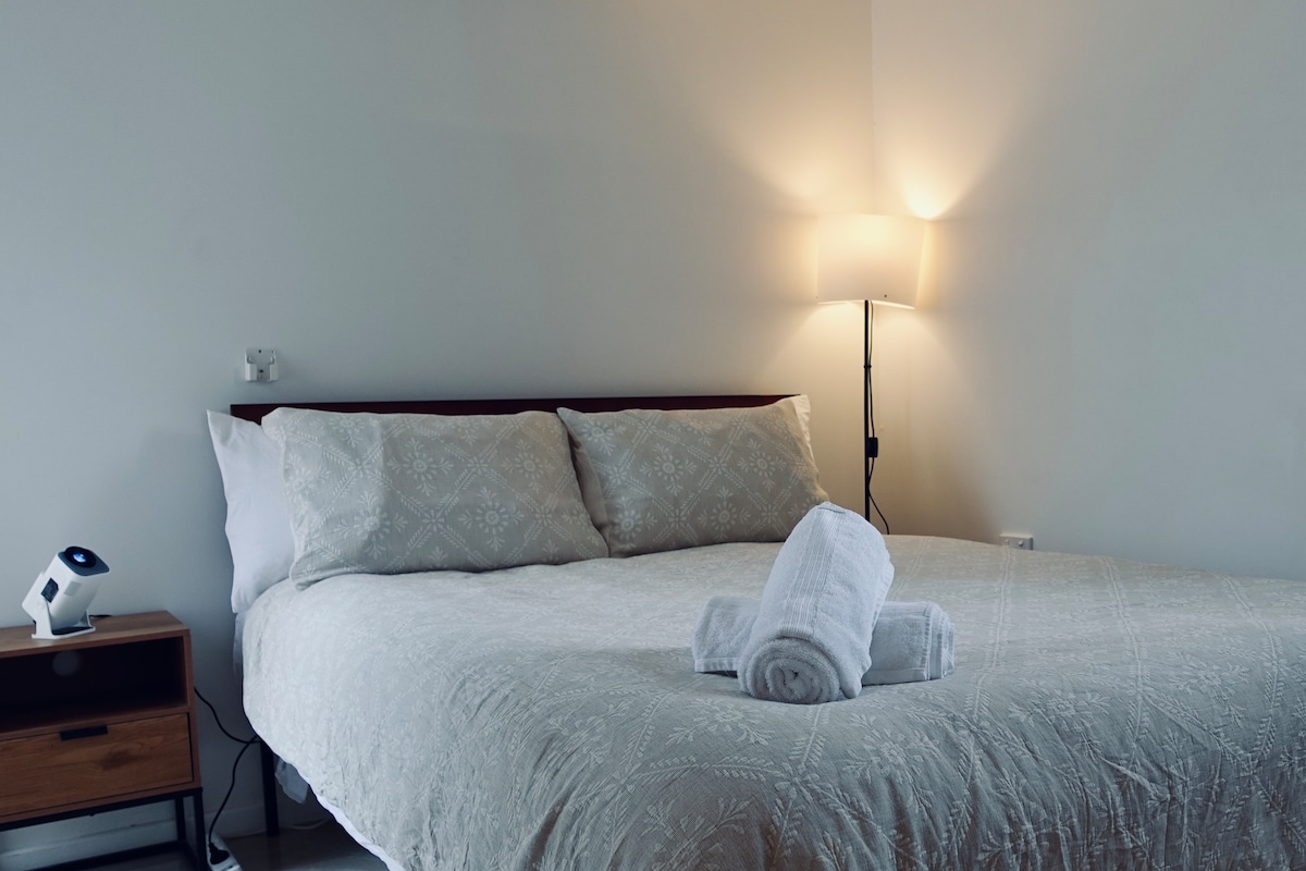 A spacious bedroom is illuminated by a warm lamp, enhancing the calm environment. The large bed is dressed in a light-coloured patterned duvet with two neatly rolled towels placed on top. A wooden nightstand is positioned next to the bed, complementing the room's simplicity.