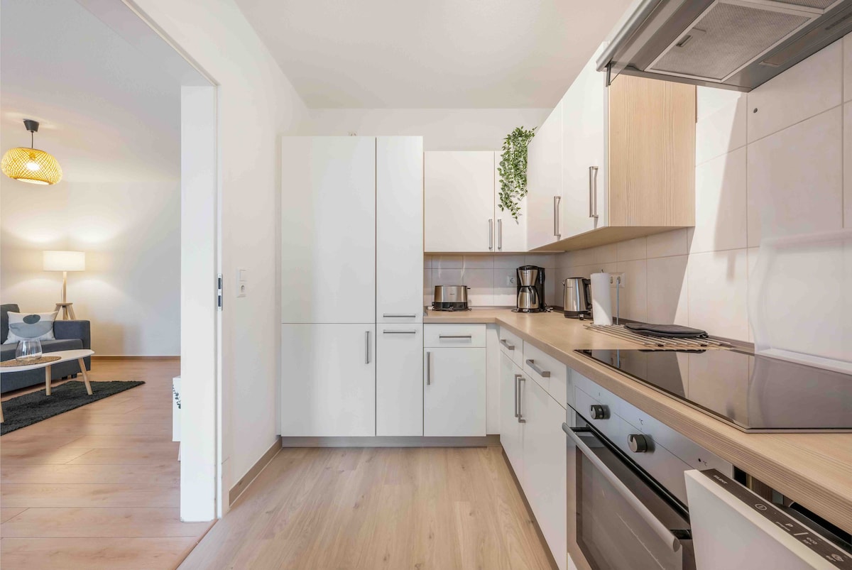 A modern kitchen is presented, featuring white cabinetry and wooden countertops. Kitchen appliances including a coffee maker and kettle are visible. The open layout flows into a living area, hinting at comfortable seating in the background. Natural light illuminates the space through an adjacent room.