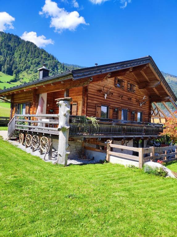 A rustic wooden chalet is showcased amid a lush green landscape. The structure features large windows, a wraparound balcony with wooden railings, and decorative elements including a wagon wheel. The scenic backdrop consists of rolling hills and a clear blue sky dotted with clouds.