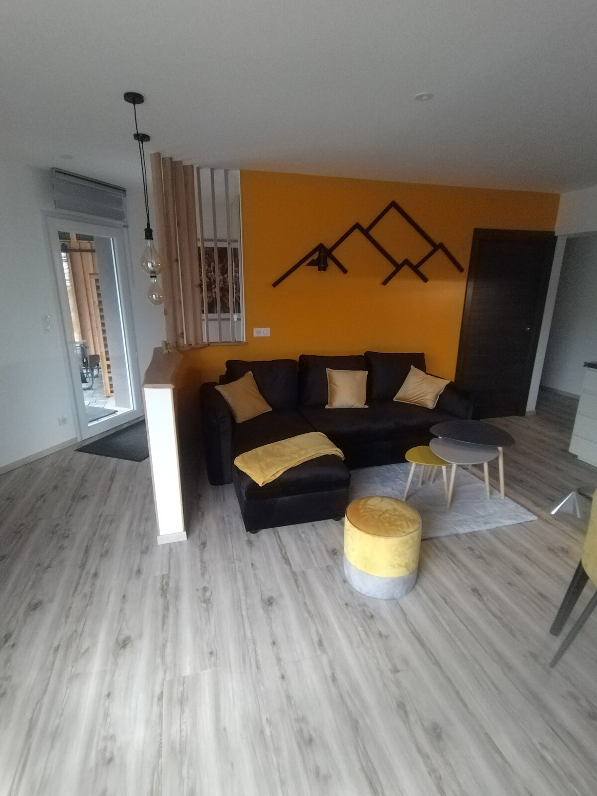 A cozy living area is presented, featuring a black sectional sofa adorned with yellow cushions. A set of small tables is positioned nearby on a textured rug. The wall is decorated with mountain-themed artwork, complementing the warm yellow accent wall. Natural light filters through a glass door.