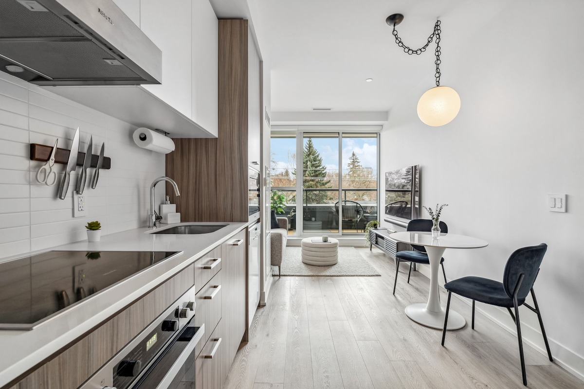 A modern kitchen area is presented with sleek cabinetry and high-end Miele appliances. A round dining table with two navy chairs is positioned beside a large window, offering natural light. A comfortable seating area with a light rug is visible, enhancing the open layout.