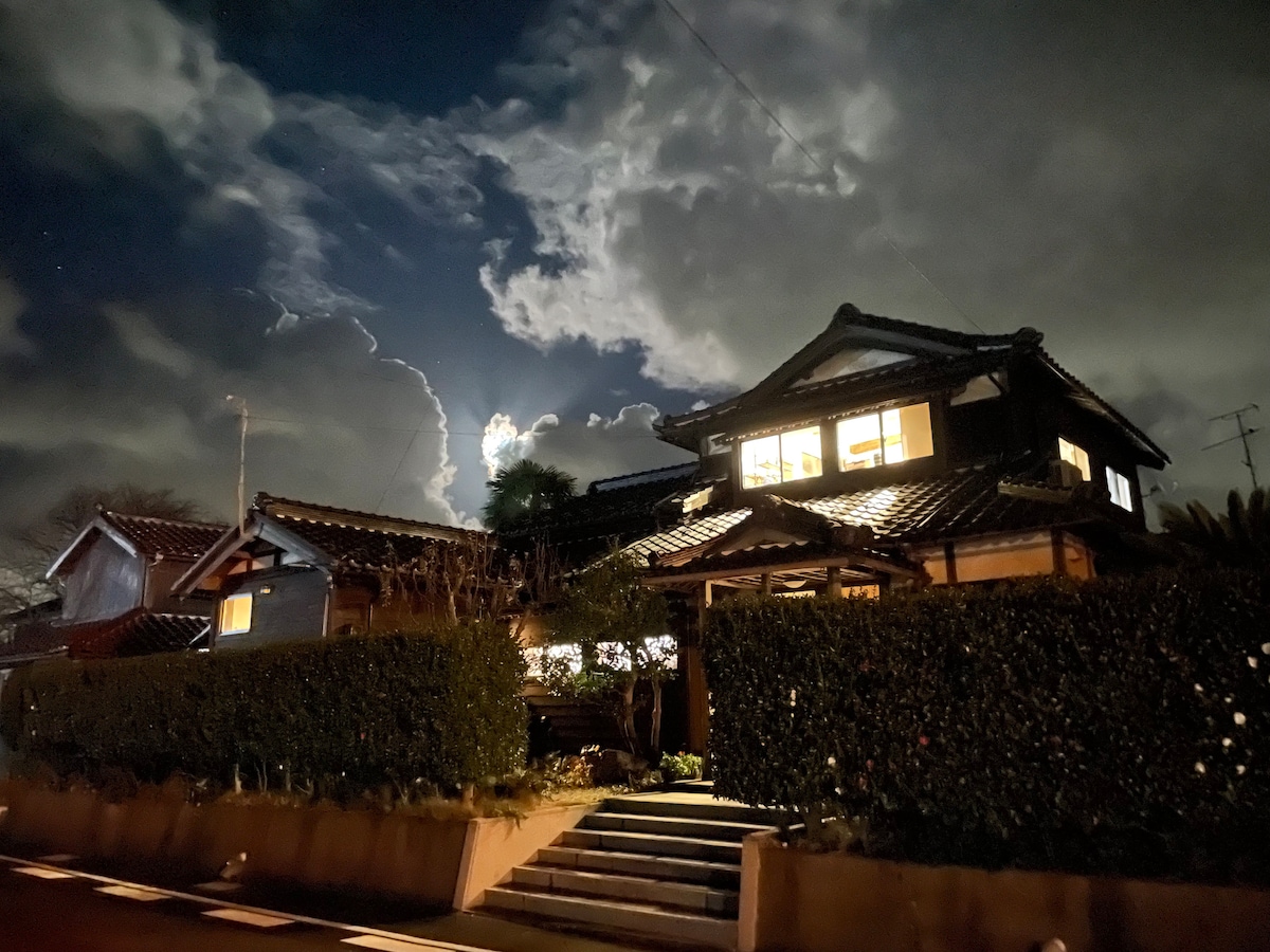 The traditional house features a sloped roof and multiple windows illuminated from within, showcasing a warm atmosphere against a dramatic night sky. Lush greenery lines the front, complemented by stone steps leading to the entrance, all framed by scattered clouds illuminated by moonlight.