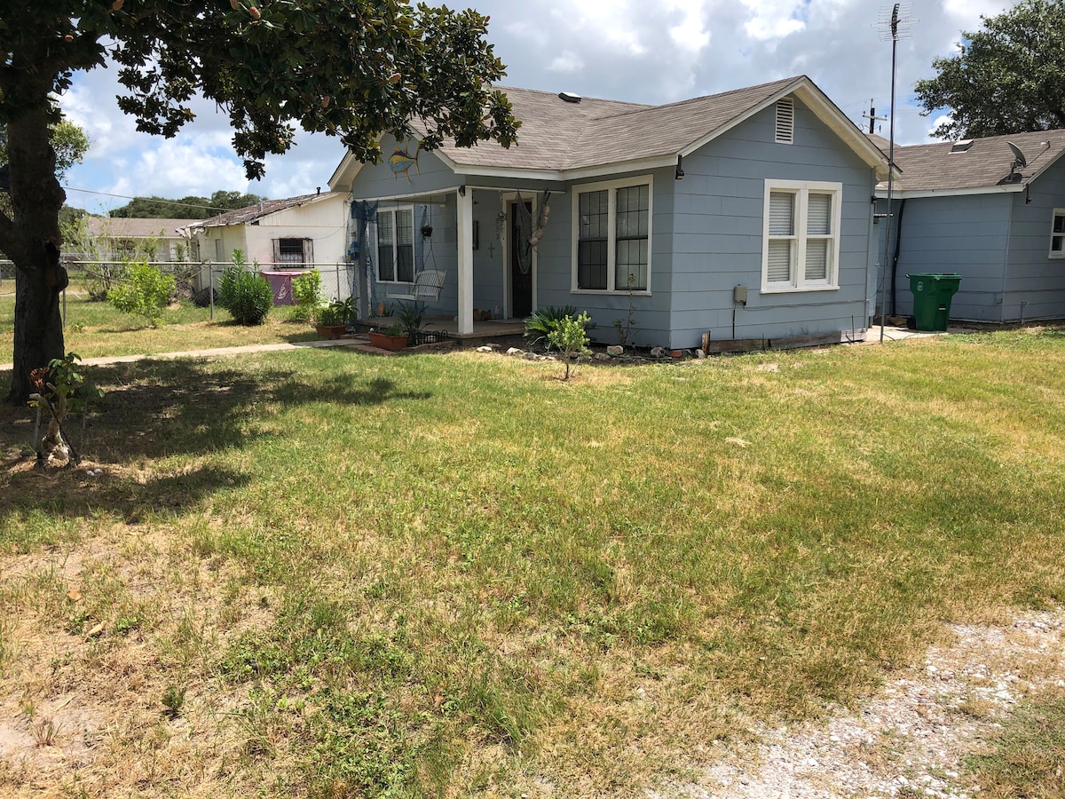 The exterior of a coastal cottage features blue siding and a well-maintained yard with grassy areas. A small porch with a chair is visible, along with a few flower beds. In the background, additional structures and a partially visible fence can be seen.