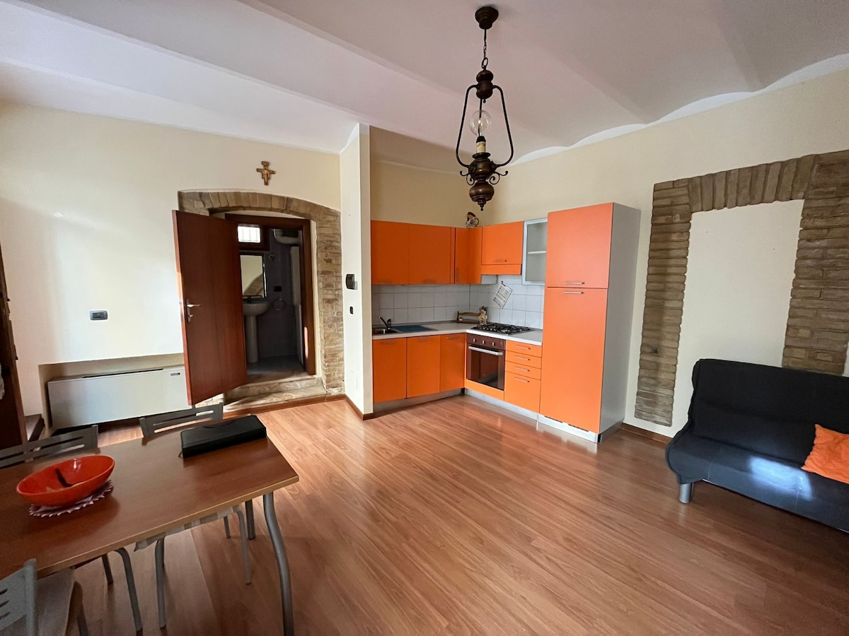 An open-plan living space features warm wooden flooring and orange cabinetry in the kitchen area. A dining table is positioned beside the kitchen, with a black sofa and decorative orange pillow in the sitting area. The space is illuminated by natural light from a window.