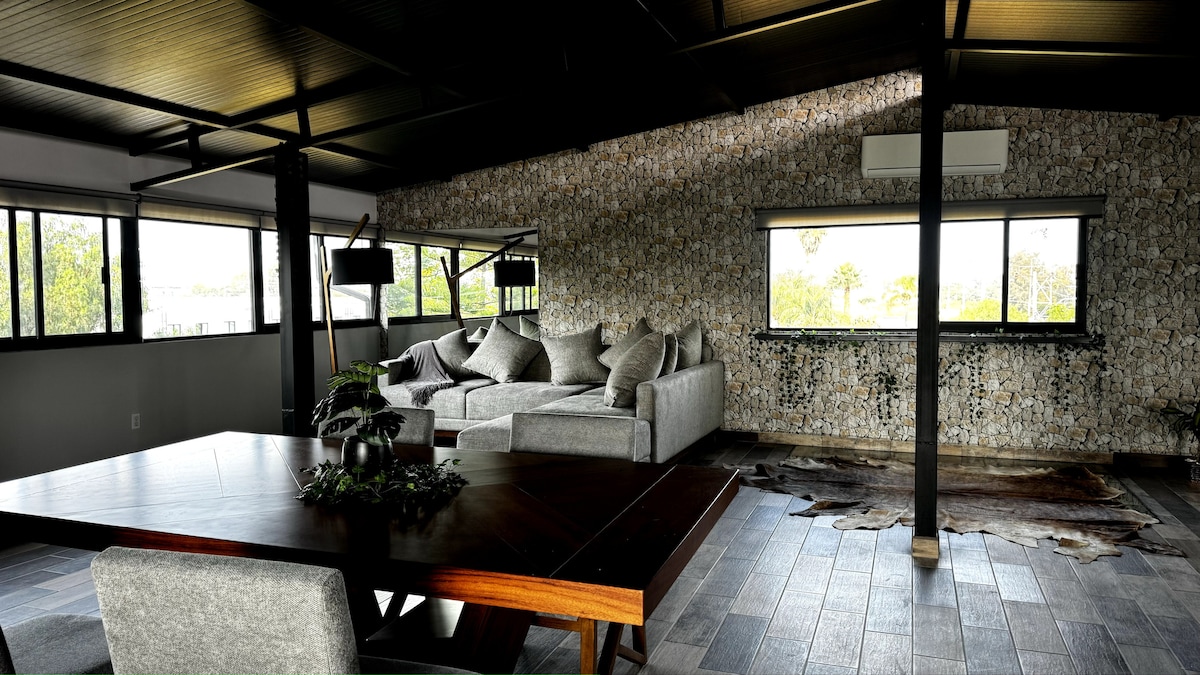 An open living space features a large sectional sofa with soft cushions, facing expansive windows that let in natural light. A solid wood dining table stands nearby, contributing to the modern decor, complemented by a feature wall adorned with natural stone accents.