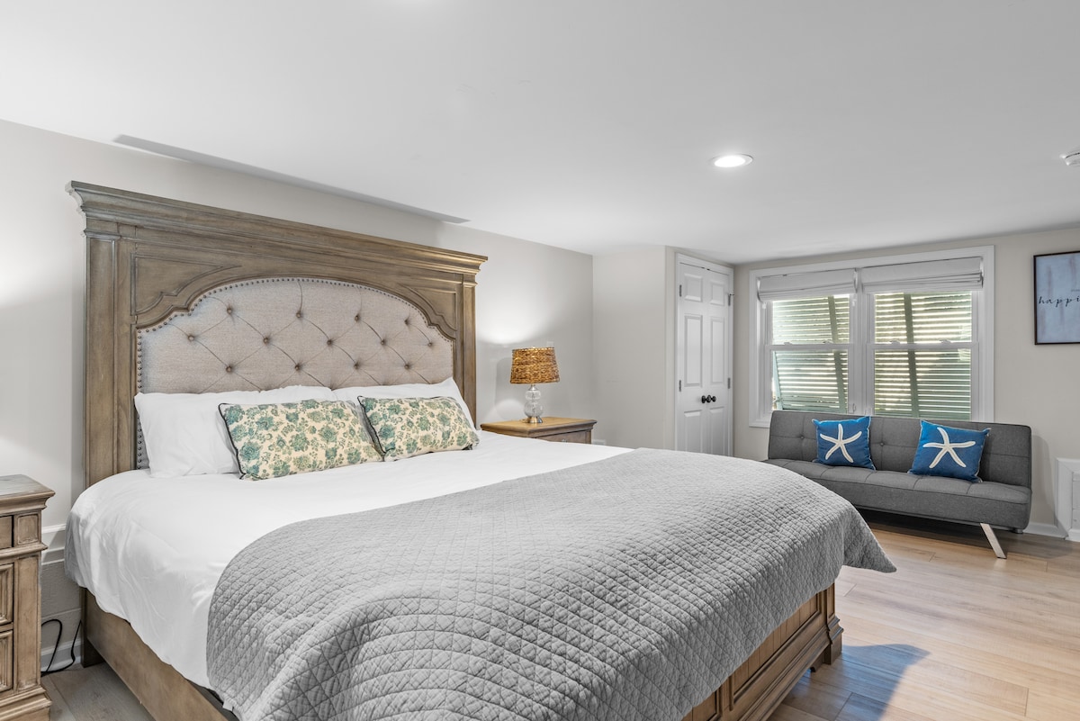 The master bedroom features a spacious king-size bed with a soft quilt and decorative pillows. A comfortable sofa is positioned nearby, complementing the natural light filtering through the window. Nightstands with lamps provide additional lighting.