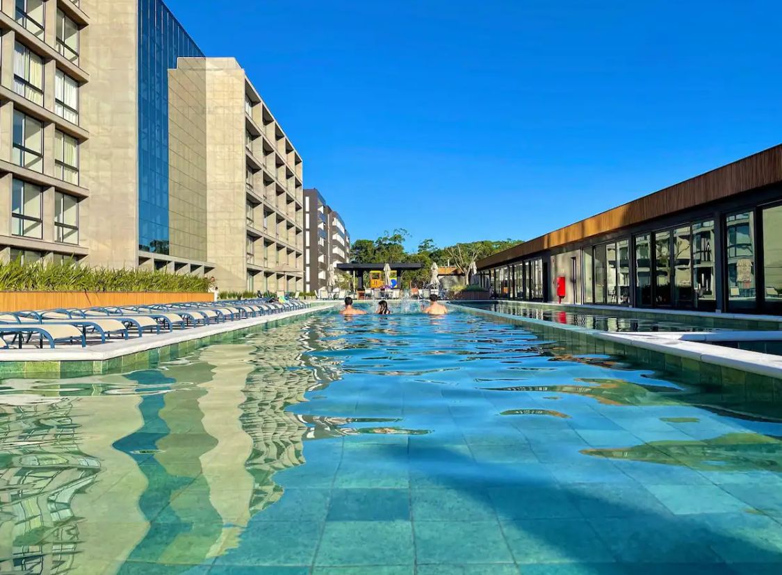 An expansive outdoor swimming pool is surrounded by lounge chairs, reflecting the clear blue sky. Modern buildings border the pool area, creating a vibrant atmosphere. The water's surface shimmers under the sunlight, inviting relaxation and leisure.