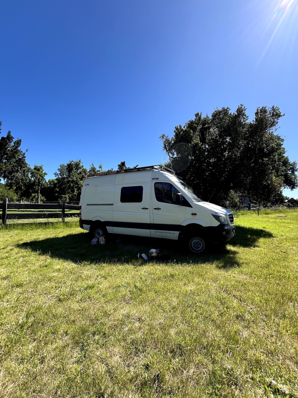 A white van is parked in a grassy area under a clear blue sky, surrounded by trees. The van's exterior is well-maintained, with the sun casting a gentle glow. The setting offers an open and natural environment, ideal for outdoor relaxation.