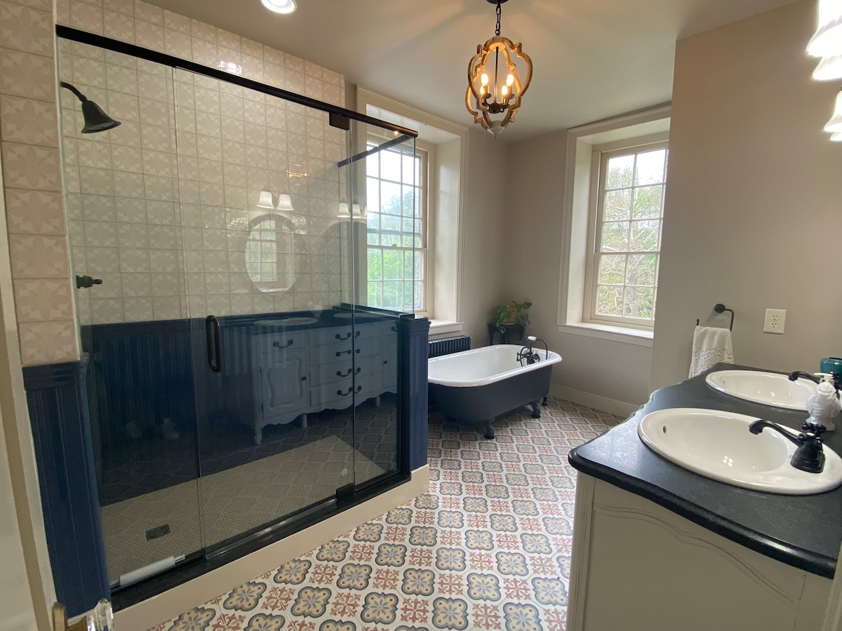 A well-appointed bathroom is featured, showcasing a glass-enclosed shower and a freestanding bathtub. Dual sinks are positioned on a dark countertop, with elegant fixtures. Natural light fills the space through two large windows, illuminating patterned tiles on the floor.