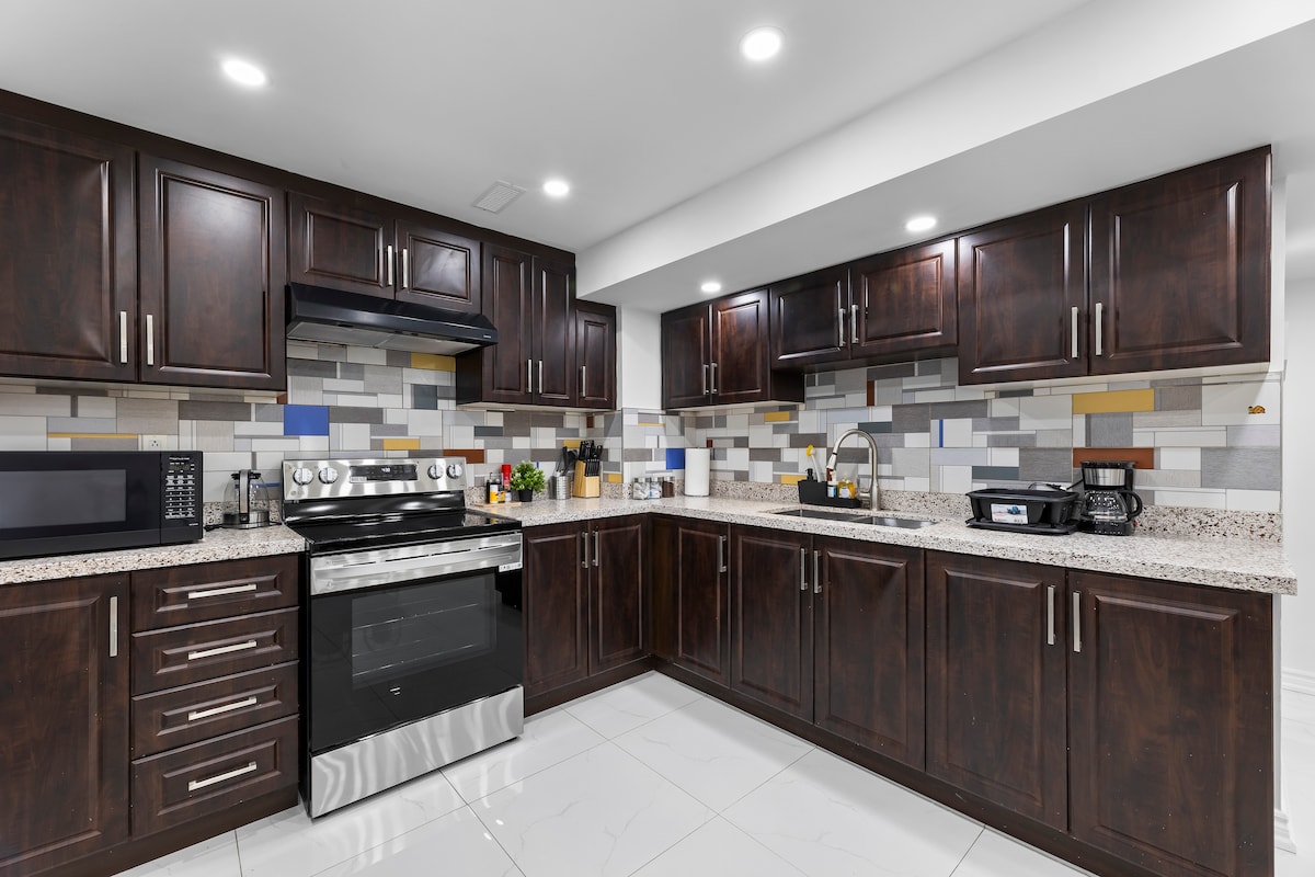 A modern kitchen is equipped with dark wood cabinetry and stainless steel appliances. The countertop features a speckled design, and the backsplash displays a mix of colorful tiles. Under-cabinet lighting enhances the workspace, complemented by various kitchen essentials and a microwave.