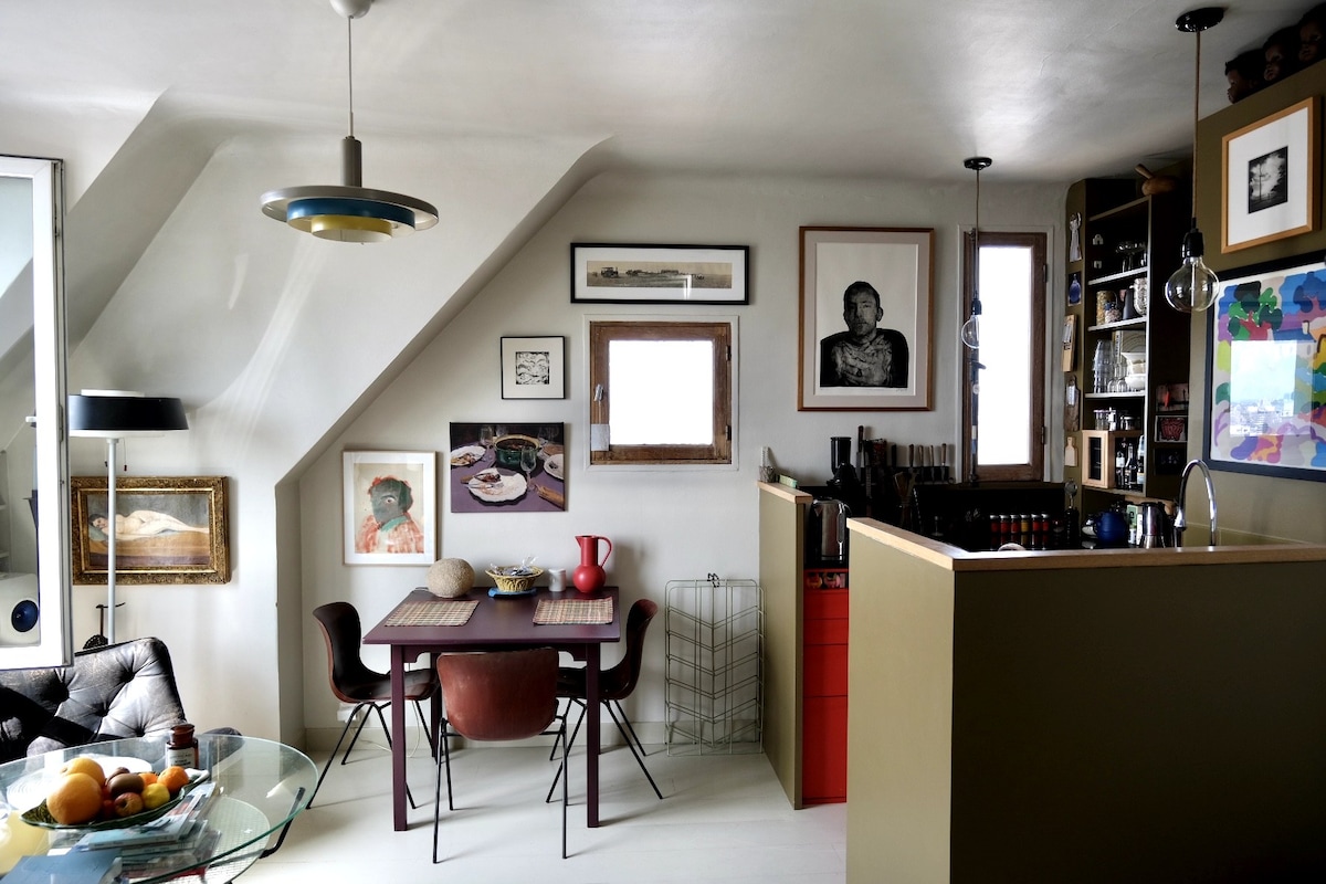 A well-appointed studio space is depicted, featuring a dining table with four chairs and a cozy seating area. Art pieces adorn the walls, complemented by natural light from windows. An open kitchen area with organized storage showcases various kitchenware, enhancing the studio's functional design.