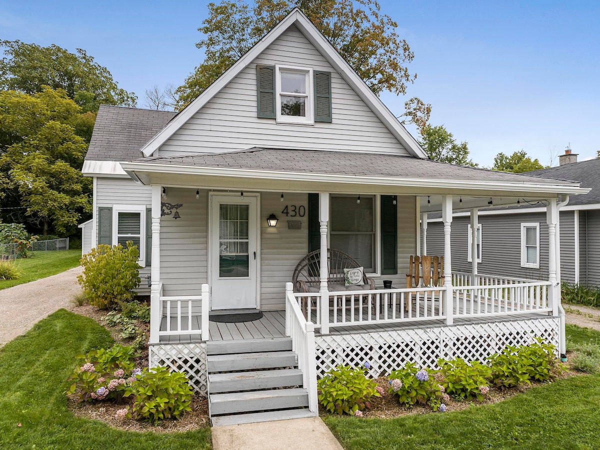 The exterior of a charming two-story house features a front porch with rocking chairs and a swing. Lush green grass and colorful flower beds surround the entrance, while trees provide a backdrop, enhancing the outdoor space's inviting quality.