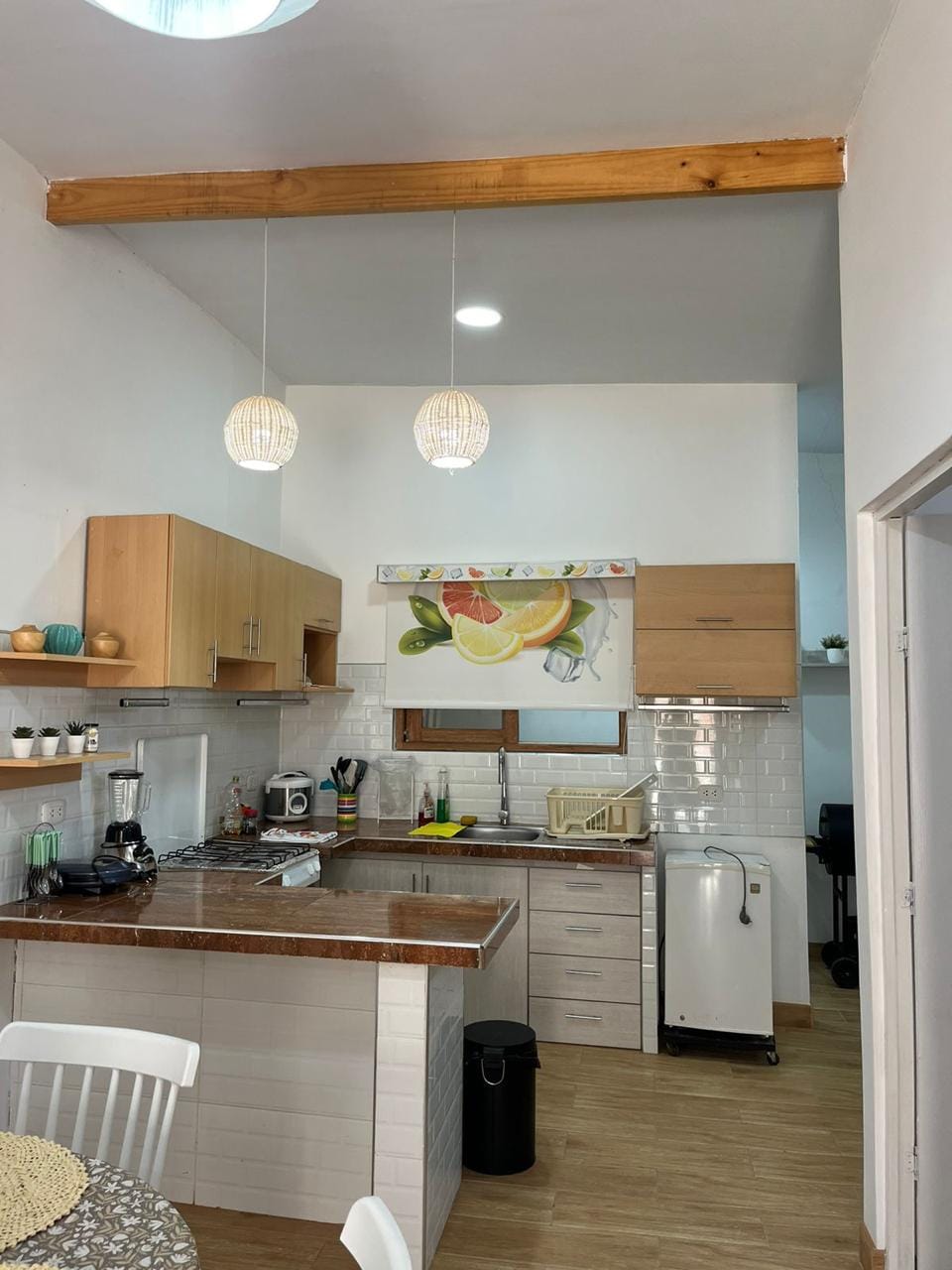 A modern kitchen features light wood cabinetry and a spacious counter. Decorative pendant lights hang above, enhancing the bright atmosphere. A wall mural of fruit adds a vibrant touch, while appliances and utensils are neatly arranged for convenience.