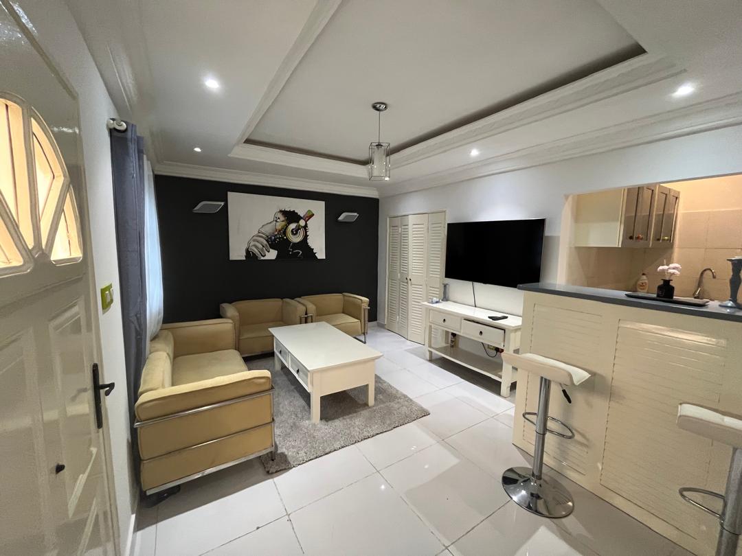 A comfortable living area features a light-colored sofa set and a coffee table situated on a soft area rug. A wall-mounted television is positioned across from the seating, with stylish wall art enhancing the decor. Kitchen bar stools are placed at a nearby counter.
