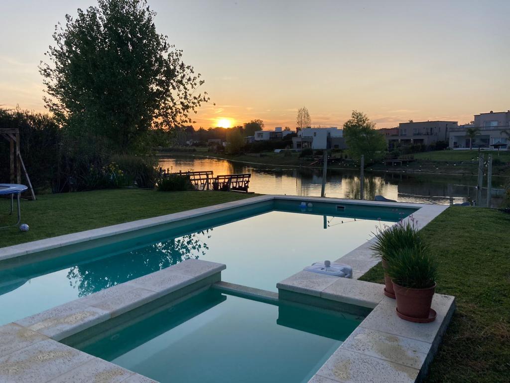 A serene outdoor pool reflects the warm hues of a sunset. Lush green grass surrounds the pool area, contrasting with the clear water. In the background, a calm lagoon is visible, framed by trees and modern homes.