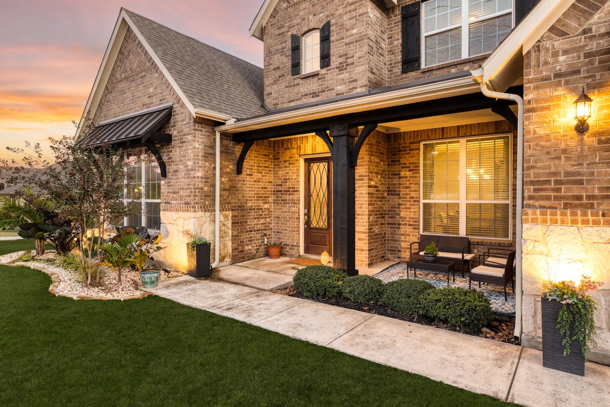 The entryway of a charming brick home features a covered porch with elegant lighting. Comfortable seating is arranged on the porch, surrounded by decorative plants and landscaping. A well-maintained lawn completes the inviting scene, illuminated by the warm glow of the setting sun.