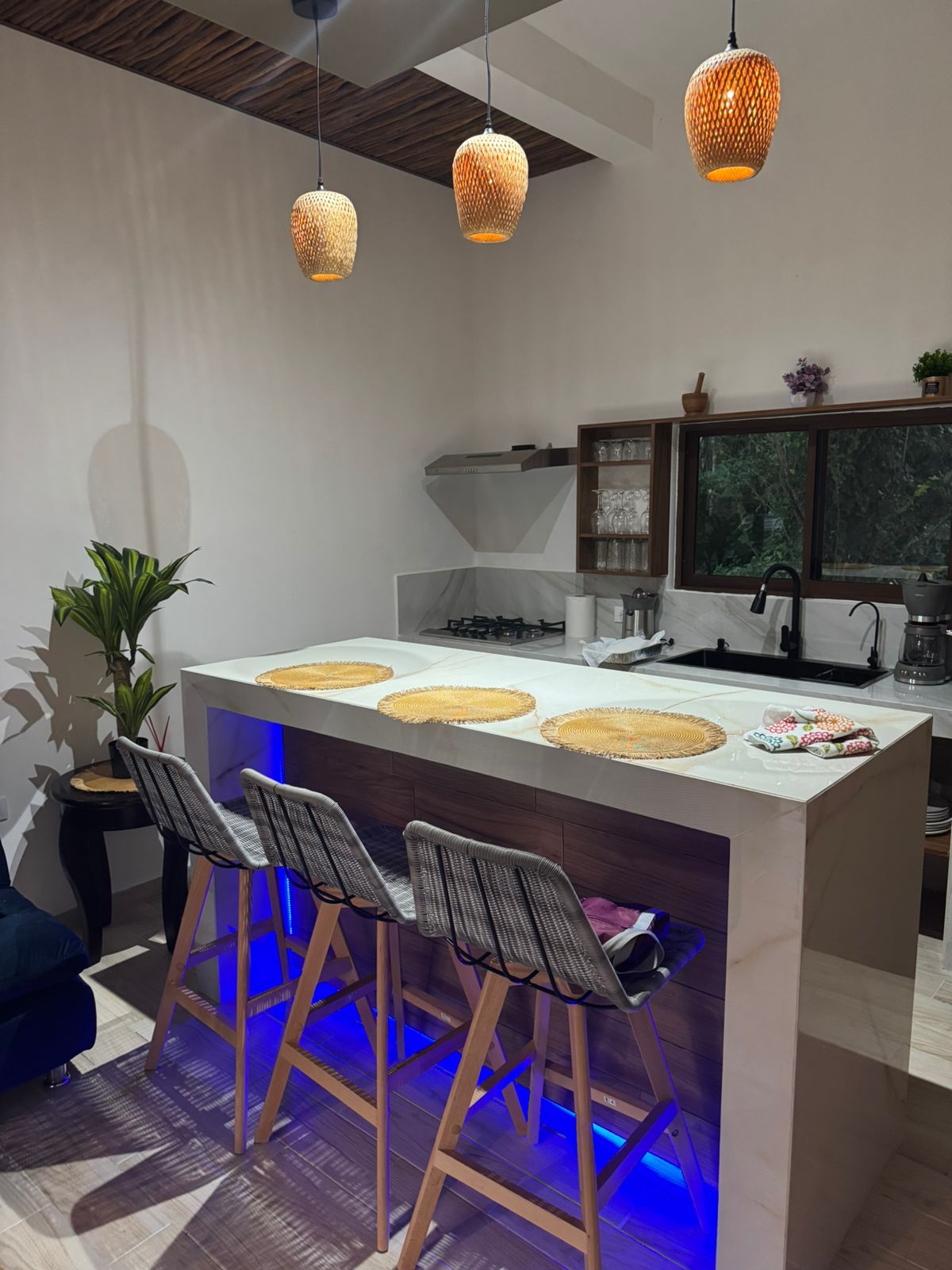 A modern kitchen is highlighted with a large island featuring three high stools. Natural light enters through a window, illuminating the space, which is accessorized with pendant lighting. A small potted plant adds a touch of greenery to the area.
