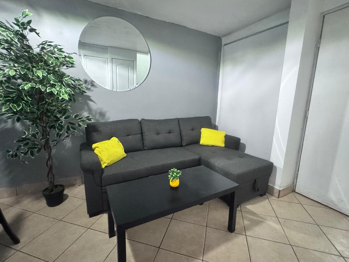 A modern living area is featured, showcasing a dark gray sofa with bright yellow cushions. A small black table sits in front of the sofa, while a decorative plant adds a touch of greenery. Light enters through a window, creating a well-lit space.