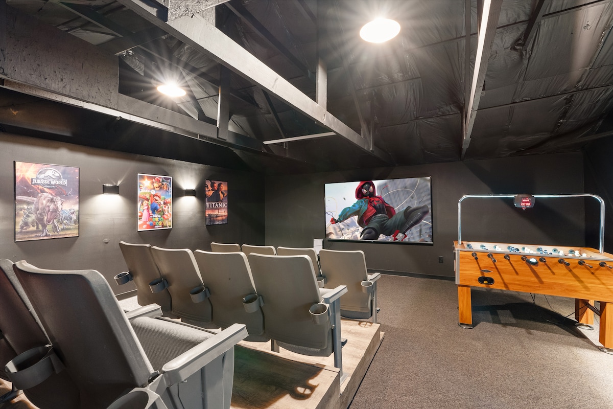 The dedicated movie theater features comfortable stadium-style seating arranged in rows, facing a large 100-inch screen. The space is accented by themed movie posters on the walls. A foosball table is positioned to one side, providing additional entertainment options.