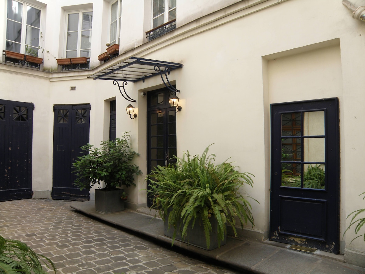 A tranquil courtyard is depicted, featuring a patterned stone pathway and greenery. Two dark-framed doors stand on either side, accented by decorative wall lanterns. Potted plants add a touch of nature, complementing the warm, muted tones of the surrounding architecture.