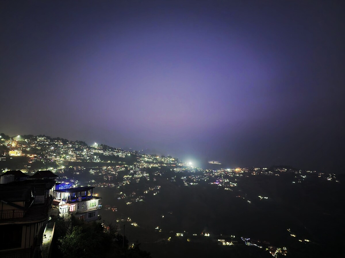 A nighttime view showcases the illuminated hills of Shimla, with twinkling lights across the landscape. The scene is enhanced by the soft glow of buildings reflecting the peaceful atmosphere of the region. The sky transitions from deep purple to darker hues, creating a serene backdrop.