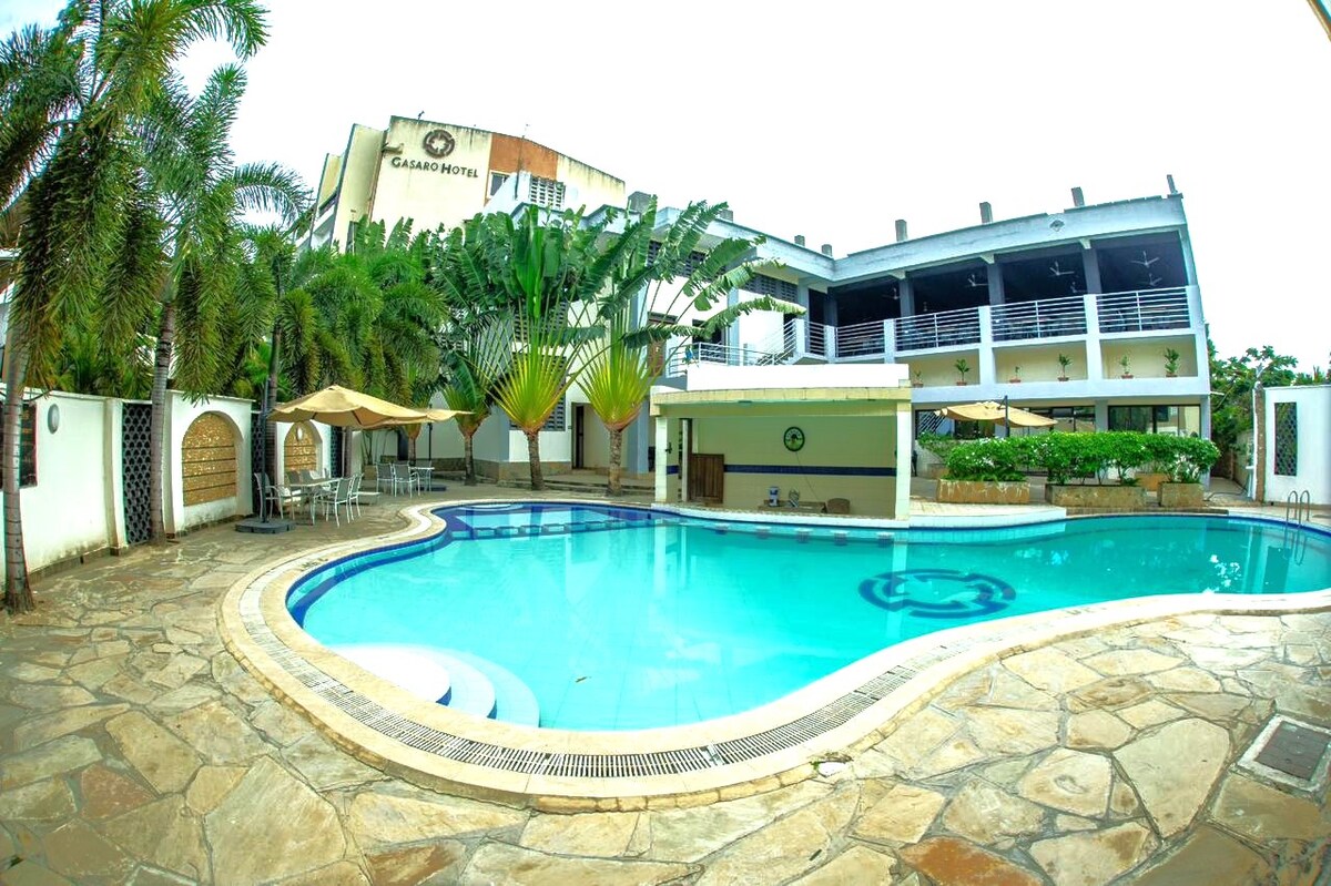 An outdoor swimming pool is surrounded by a stone patio and lush greenery. Sun loungers with umbrellas are positioned around the pool, and a restaurant terrace overlooks the water. The Gasaro Hotel building is visible in the background, featuring balcony seating and multiple levels.