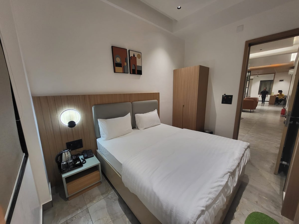 A well-appointed room features a spacious bed with neatly arranged white bedding. The contemporary decor includes a wooden accent wall, bedside lights, and minimalistic artwork. A wardrobe is present alongside a small side table holding a coffee maker, creating a functional and inviting space.