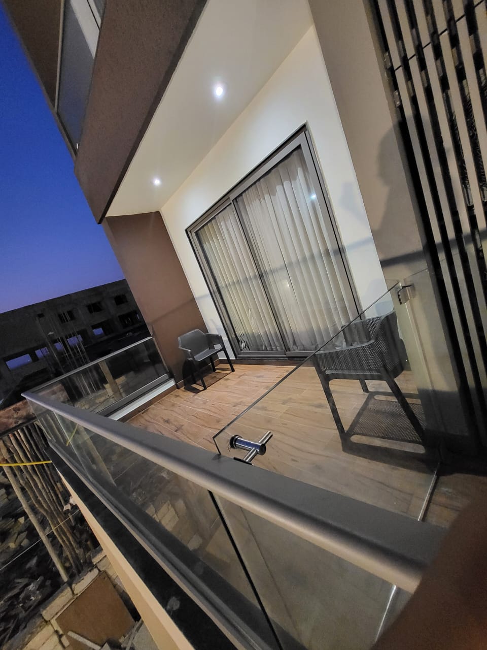 A private balcony is shown with two black wicker chairs positioned next to a glass railing. Sliding glass doors allow access to the interior space. The wooden deck adds warmth, and the evening sky is visible, transitioning to twilight.