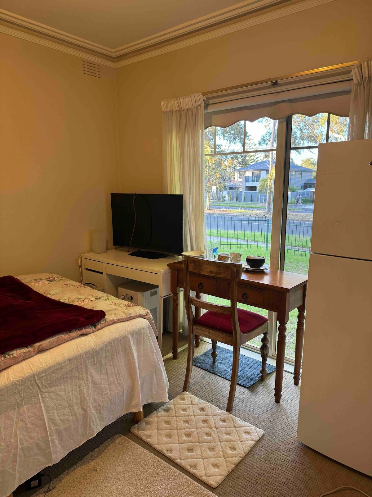 A cozy room features a double bed with a soft white coverlet. A small wooden desk with a chair is positioned next to a television. Natural light enters through a large window, offering a view of the outside area. A refrigerator stands nearby.