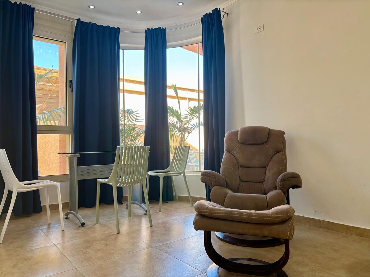 A bright and airy corner nook features a comfortable rocking chair next to a small glass table. Two light-colored chairs are positioned nearby, with large windows allowing natural light to fill the space, complemented by dark blue curtains.