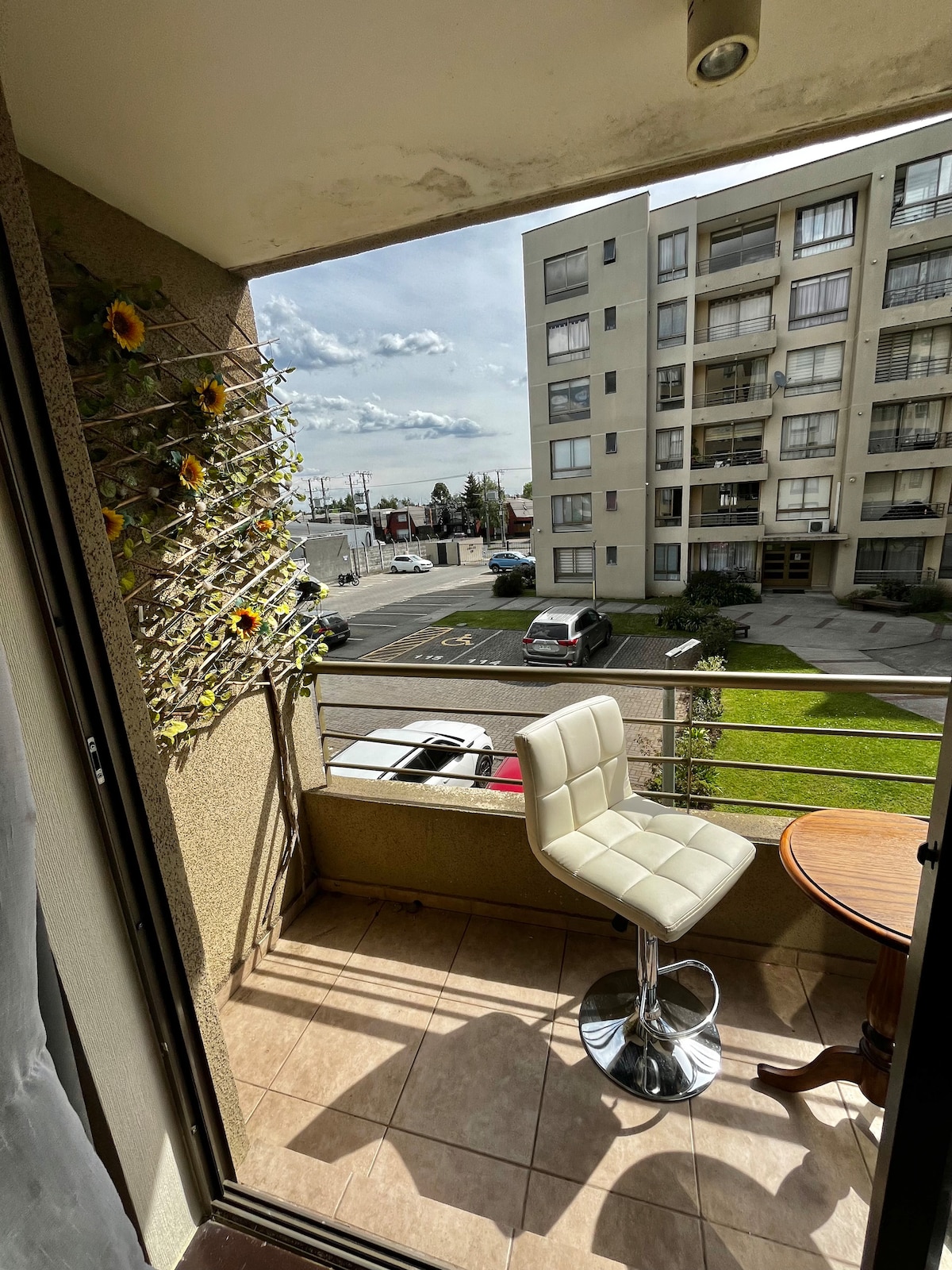 A small balcony space is depicted, featuring a round wooden table paired with a modern white barstool. A trellis adorned with vibrant green leaves and sunflowers provides a touch of nature, while a view of the surrounding area is visible beyond the railing.