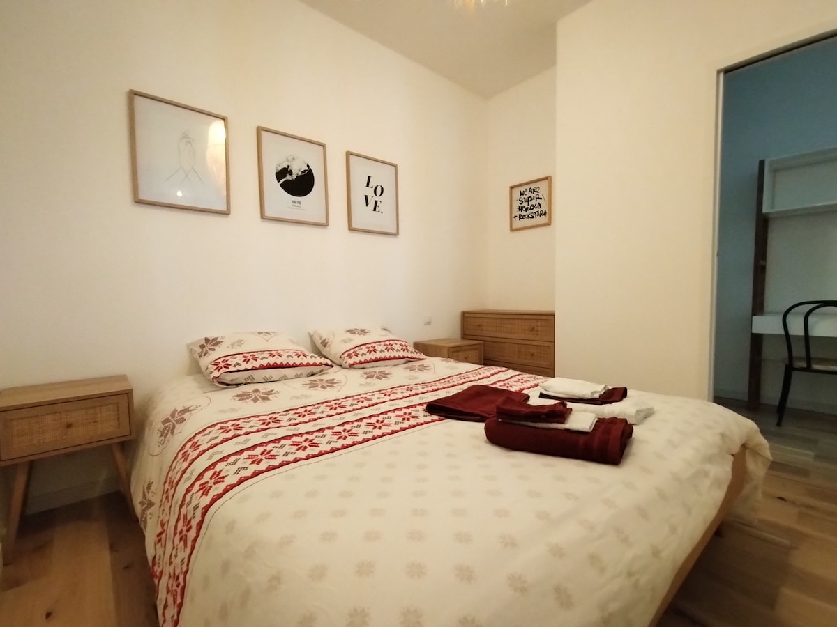 A cozy bedroom features a spacious bed with patterned bedding. Framed artwork is displayed on the walls, adding a touch of decor. Nightstands are positioned on either side of the bed, and natural light fills the room through an open door leading to another space.