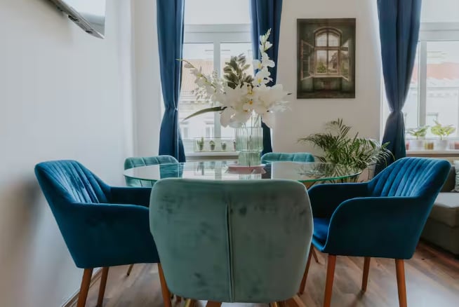 A bright dining area features a round glass table surrounded by four plush blue chairs. Two large windows allow natural light to fill the space, while a decorative vase with white flowers sits at the center. A green potted plant is visible in the background.