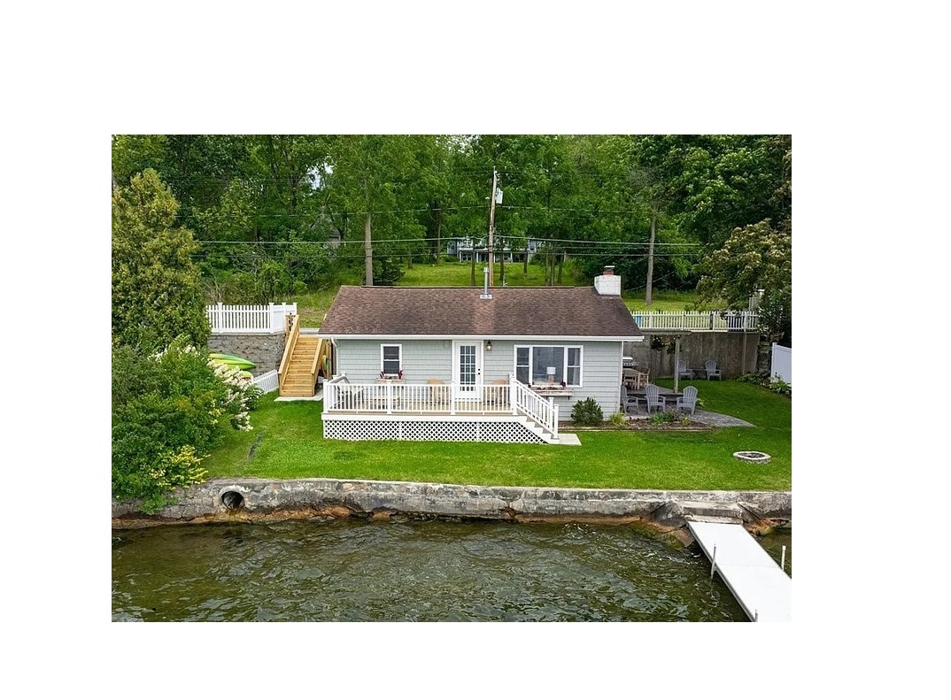 A quaint cottage is positioned near the water's edge, featuring a spacious deck surrounded by lush greenery. A gentle slope leads up to the entrance, and a dock extends into the lake. The structure is complemented by a manicured lawn and nearby landscaping.