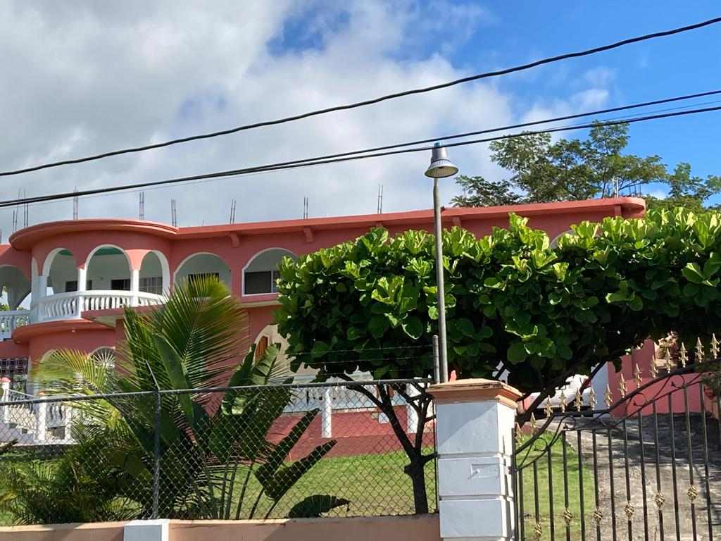 The exterior of the property showcases a colorful two-story building with arched windows and balconies. Lush green foliage and a palm tree are visible in the foreground, adding to the tropical ambiance. Clear blue skies create a bright backdrop.
