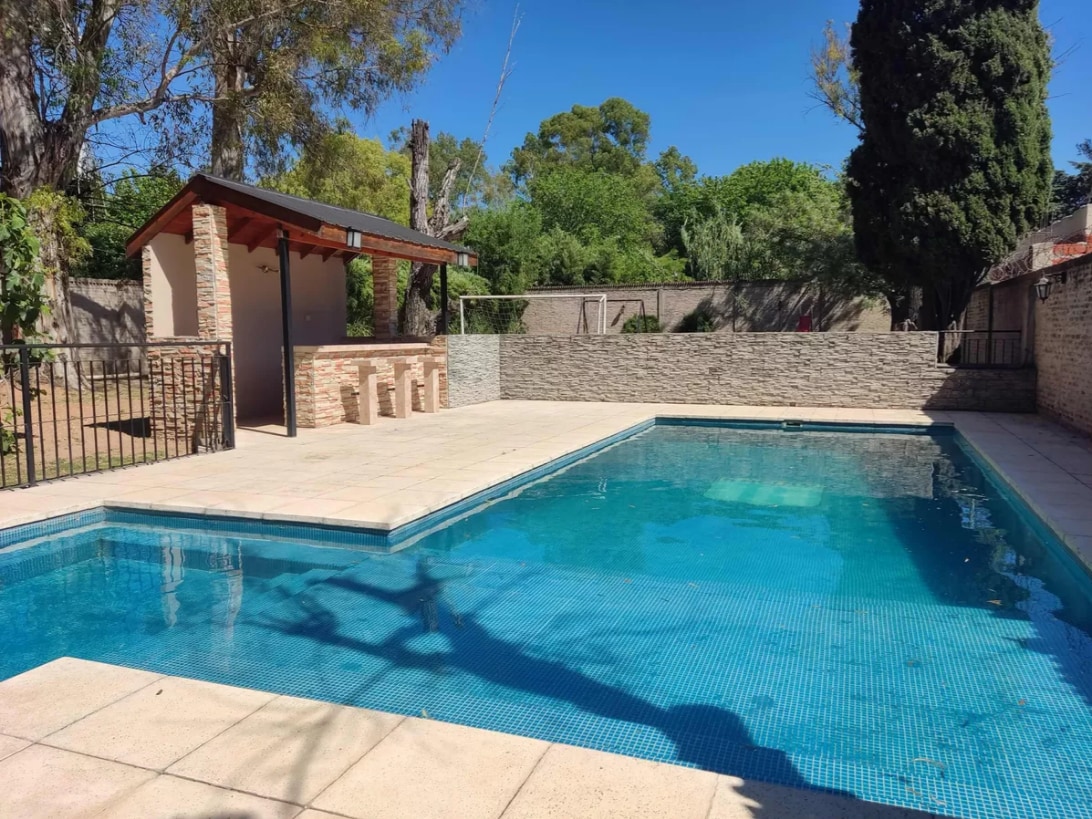 A swimming pool is surrounded by a sunlit patio, featuring clear blue water. A shaded area with a roof offers a space for relaxation, while stone walls and lush greenery frame the scene, contributing to a serene outdoor atmosphere.