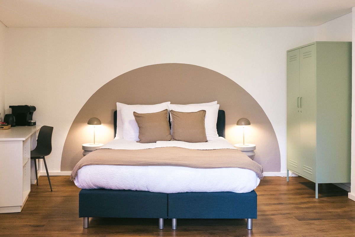 A spacious room features a generous double bed with neutral-toned bedding and accent pillows. Soft lighting is provided by two bedside lamps. A small work area includes a desk and a chair, with a kitchenette visible in the corner. The flooring is a warm wood texture.