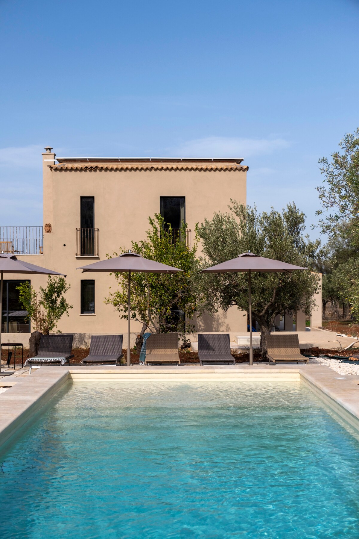 A serene outdoor area features a private pool bordered by comfortable loungers under large umbrellas. Olive trees provide natural shade, with a rustic building in the background enhancing the peaceful setting. The clear blue water of the pool contrasts beautifully with the landscape.