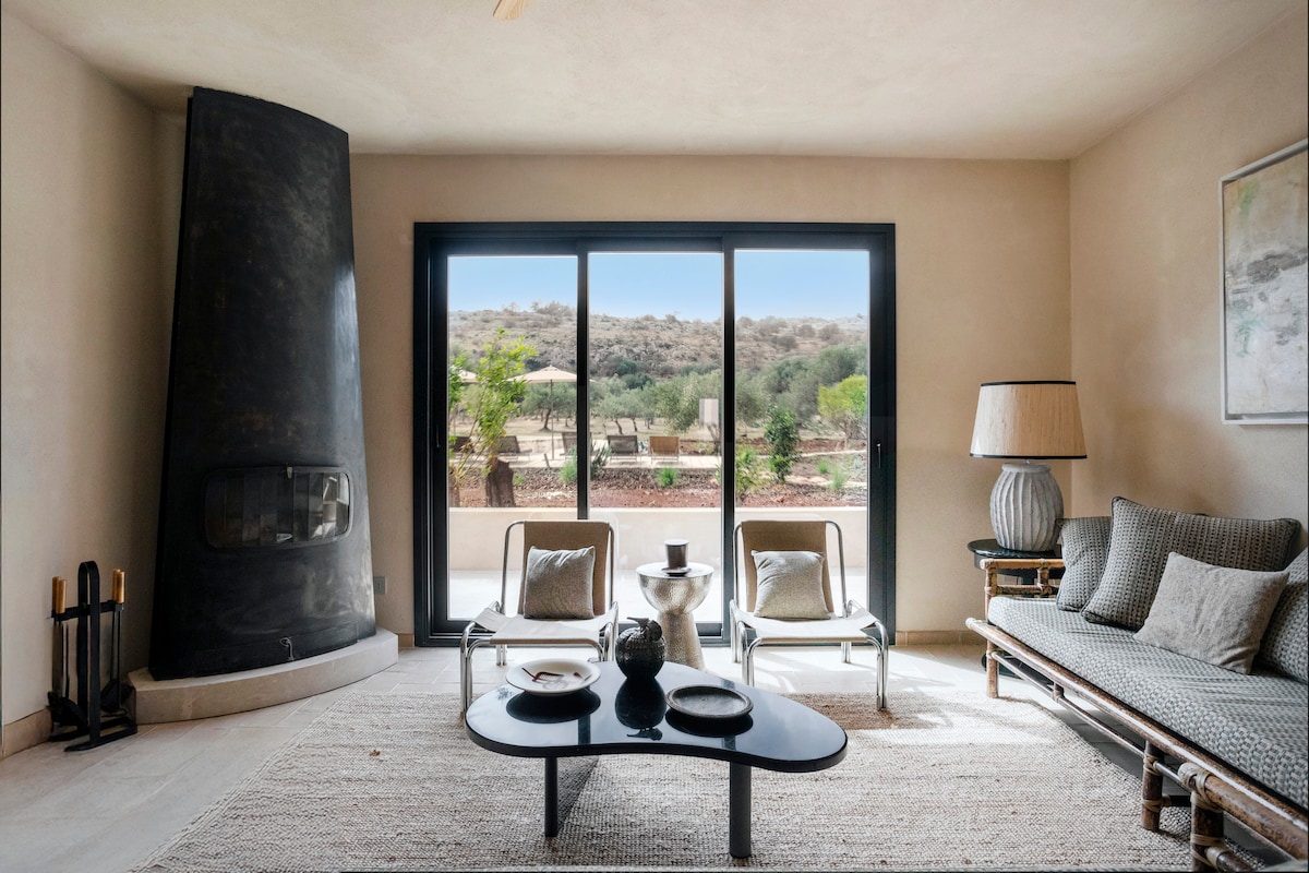 A modern living area features a sleek fireplace and large glass doors that open to a scenic outdoor view. A comfortable sofa and two chairs surround a round coffee table, creating a relaxed space for gatherings. Natural light enhances the soft neutral tones of the room.