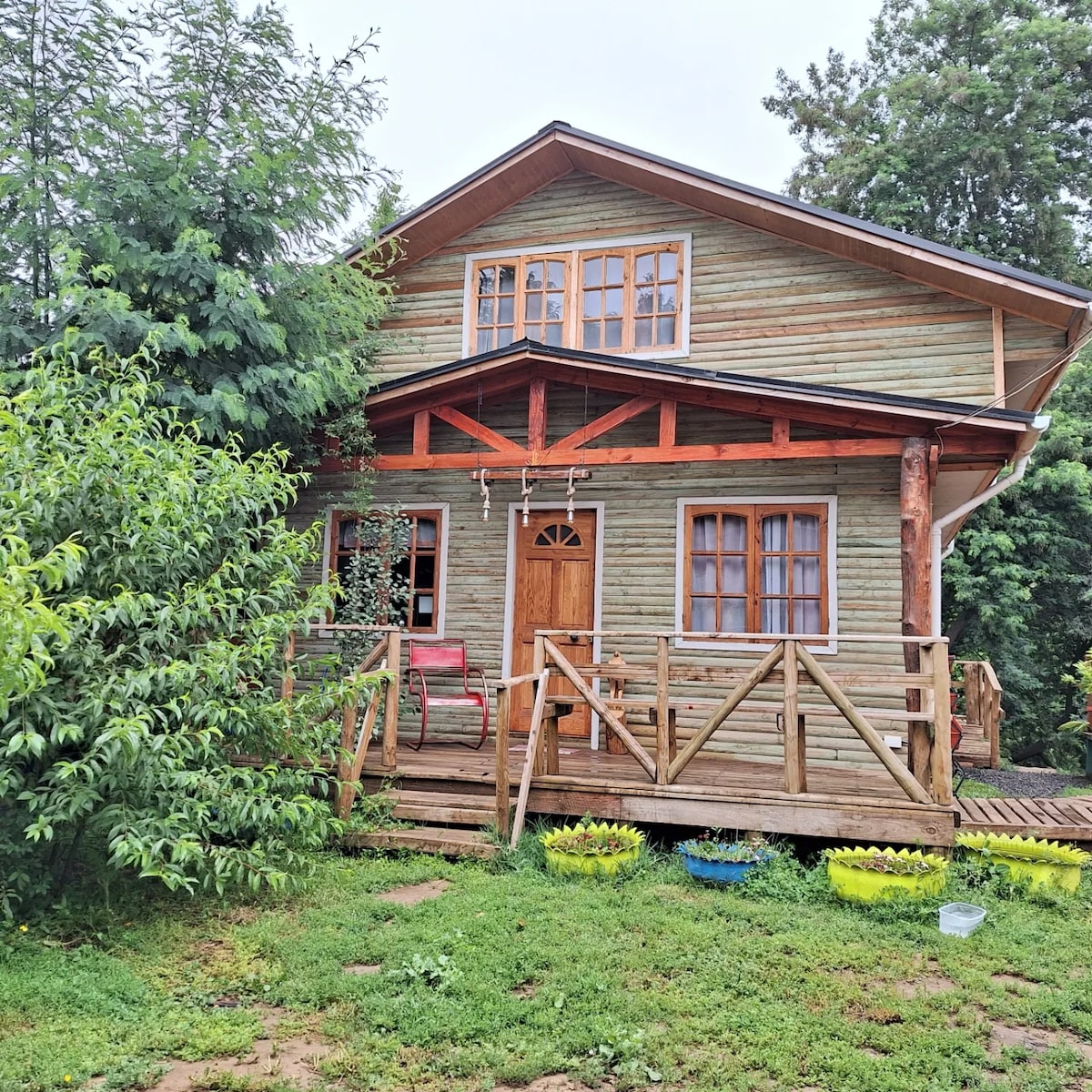A two-story wooden cabin is surrounded by greenery, showcasing a welcoming front porch with wooden railings. Large windows provide natural light, and a bright red chair is positioned near the entrance. The lush lawn features various planted pots, enhancing the natural setting.