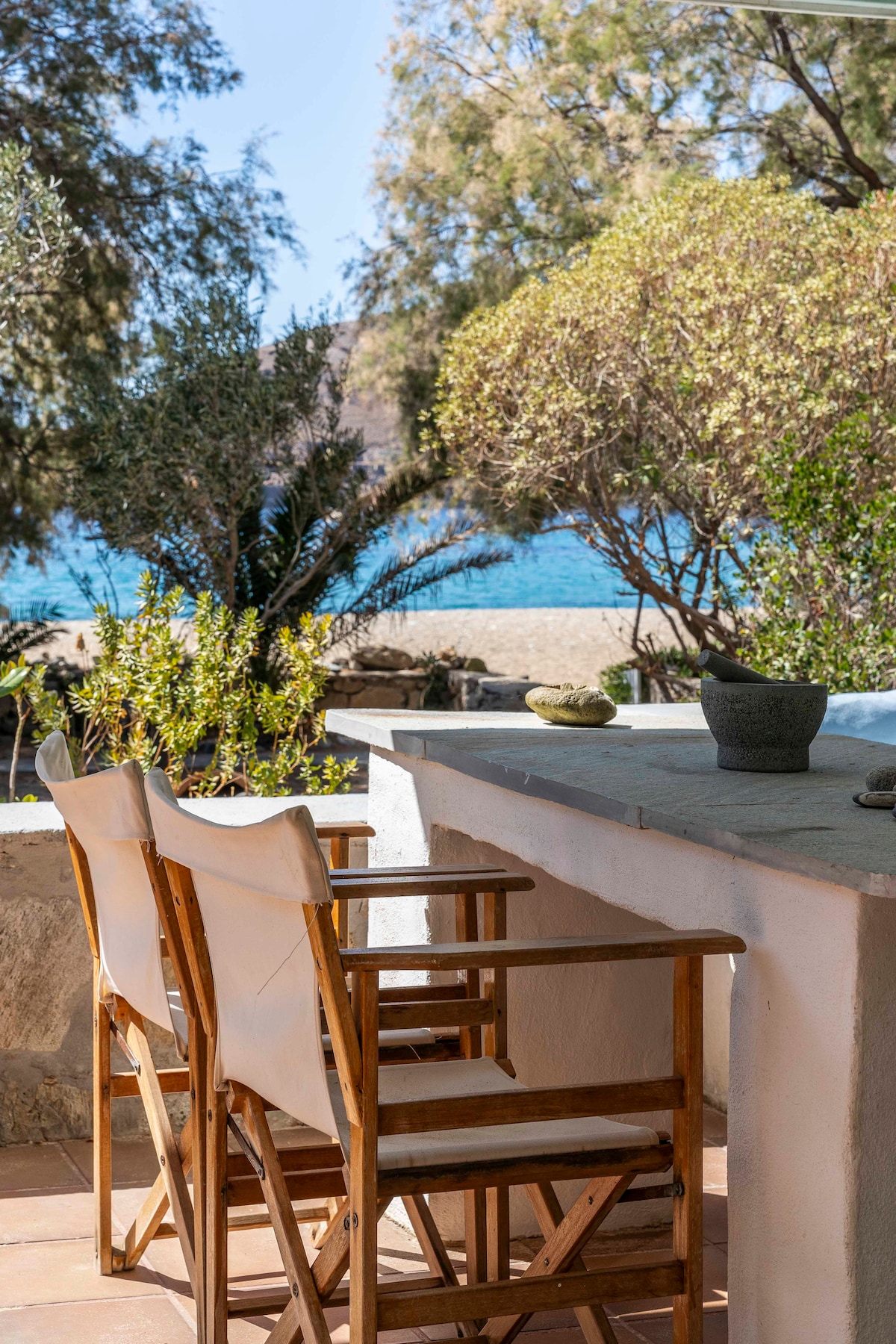 A tranquil outdoor seating area features two wooden chairs placed next to a stone countertop. Lush greenery surrounds the space, offering a peaceful ambiance. The blue sea can be seen in the background, with natural light enhancing the serene environment.