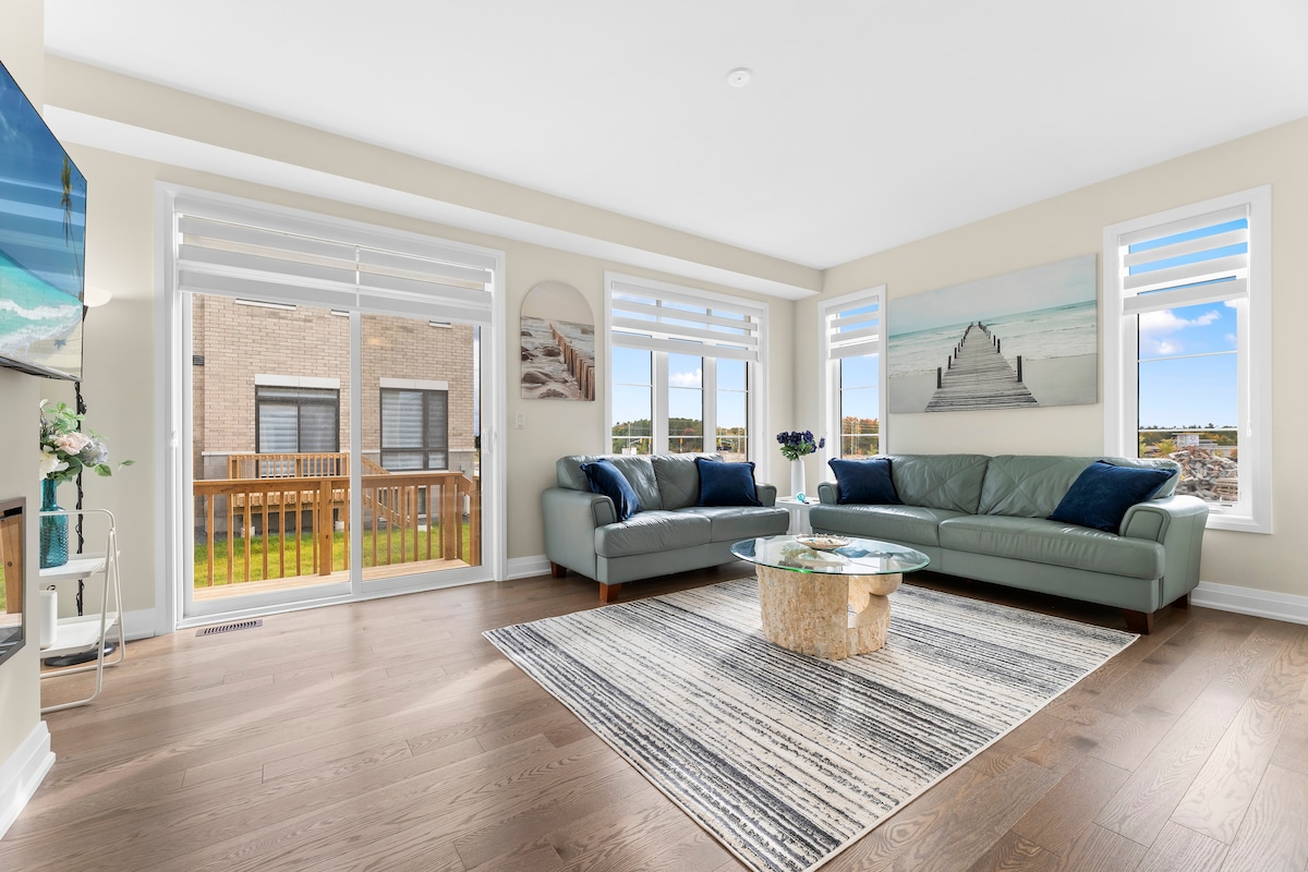 A bright living area features two comfortable sofas in soft tones, arranged around a glass coffee table. Floor-to-ceiling windows allow natural light to fill the space, with views of the balcony and outdoor scenery. The room is accented by a beach-inspired art piece above a large area rug.