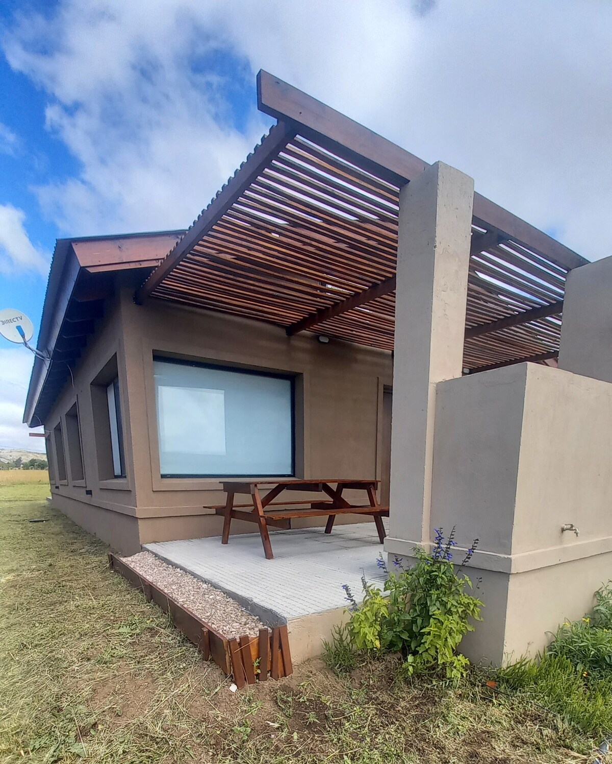 An exterior view of the duplex cabin showcases a covered patio with wooden slats, providing shade. A picnic table is positioned on a stone surface, surrounded by greenery. Large windows allow natural light into the interior, set against a backdrop of open fields and a cloudy sky.