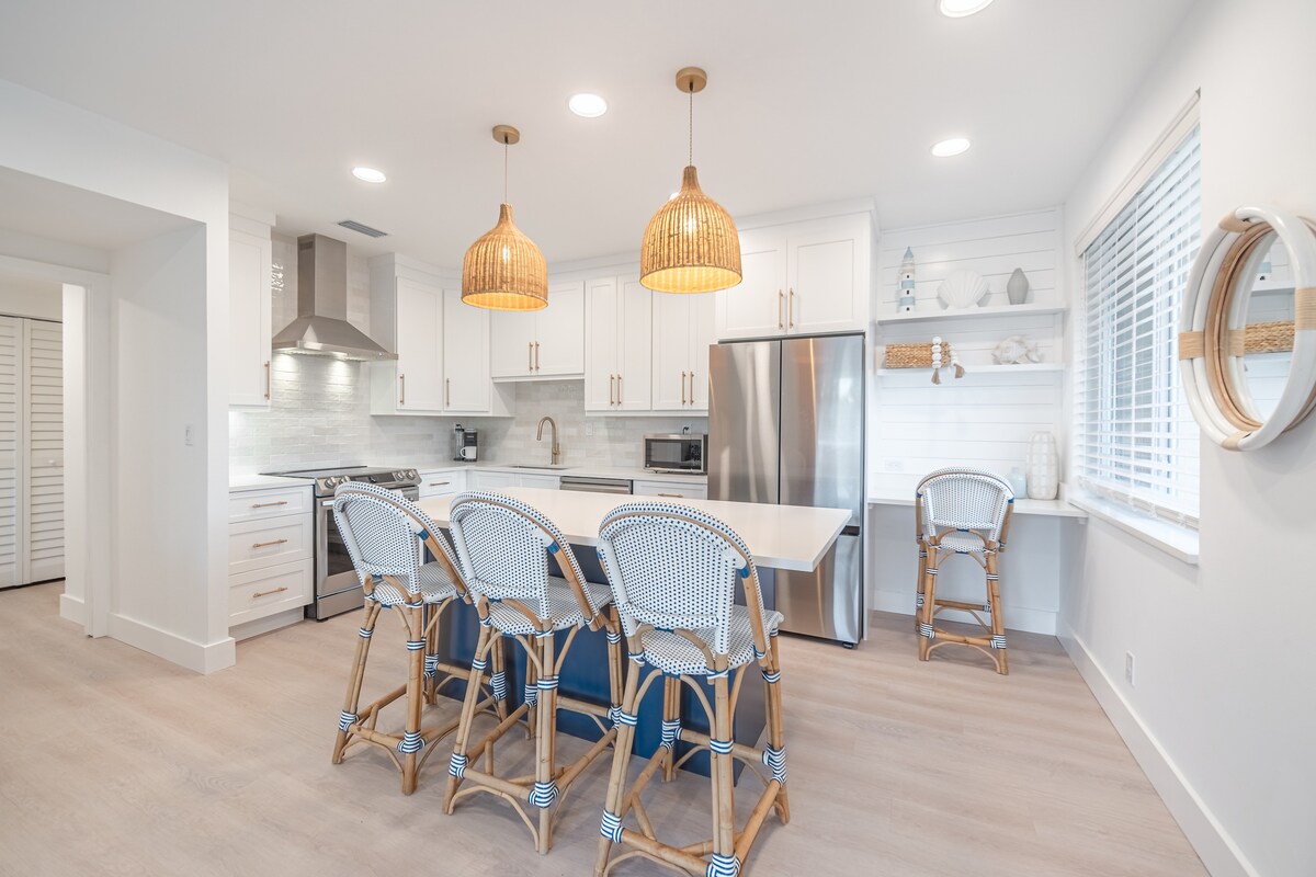 A modern kitchen equipped with high-end stainless steel appliances and custom cabinetry. Two rattan pendant lights hang over a spacious island with seating for four. Light wood flooring complements the bright white walls, enhancing the open and airy atmosphere.