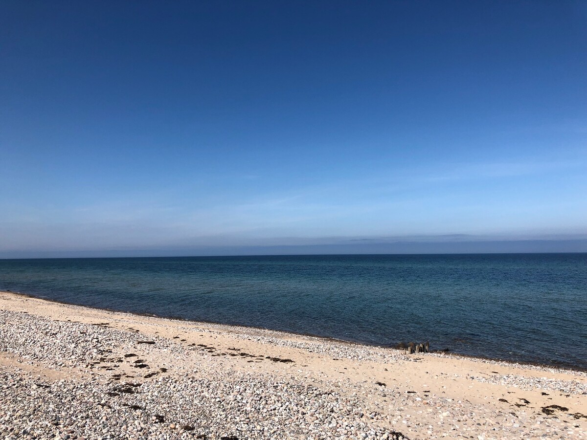 A sandy beach extends along the shoreline, bordered by smooth pebbles. The clear blue sea meets a vast sky, illuminated by natural light. Gentle waves can be seen lapping at the shore, creating a serene coastal atmosphere.